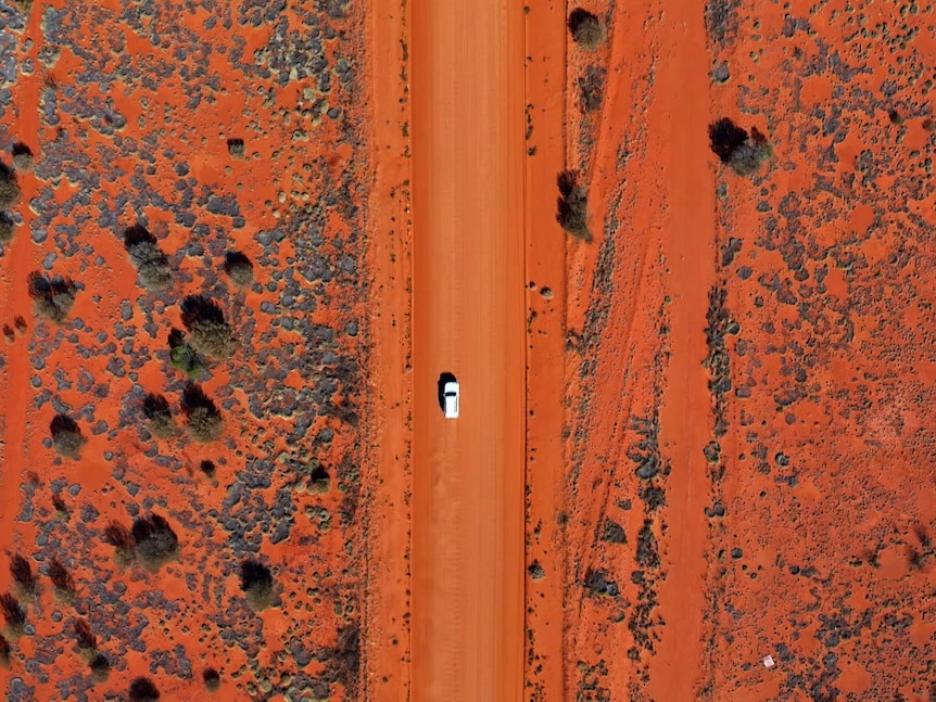 An aerial view of a white car on a red dirt highway. Either side of the road is arid desert with sparse greenery