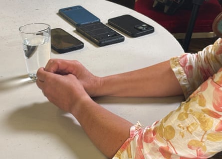 A view of a woman’s arms as she sits at a table speaking to reporters, whose smartphones recording the conversation are visible