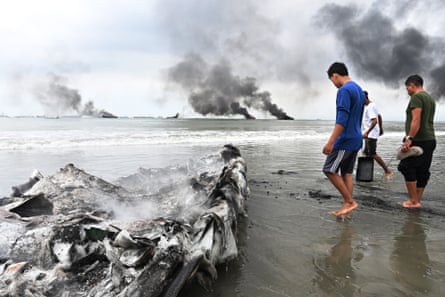 People walk on sand near water with smoke plumes in the background and a charred boat nearby