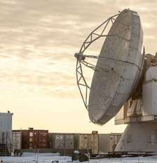 A Base Espacial Pituffik (antiga Base Aérea de Thule) é mostrada no norte da Groenlândia, em 4 de outubro de 2023. A imagem mostra uma grande antena parabólica em frente a contêineres, com um céu alaranjado e nublado ao fundo. Foto: Thomas Traasdahl/Ritzau Scanpix/AFP via Getty Images / BBC News Brasil