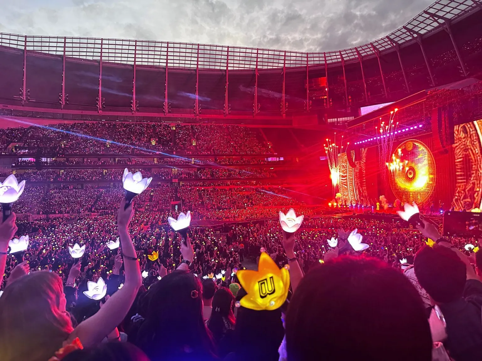 Crowd members in the foreground hold illuminated crown-shaped light sticks, including a yellow light stick with a black logo. Thousands of spectators fill a large stadium, many holding similar white glowing light sticks. On the right, a concert stage is lit in red and orange, with a large circular screen, bright stage lighting and pyrotechnic effects. The stadium seating rises around the arena, and a cloudy evening sky is visible above the open roof structure.
