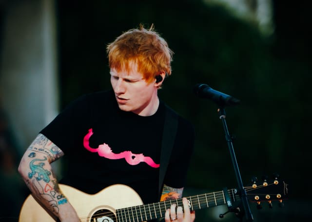 Ed Sheeran, wearing a black t-shirt with reads 'Loop', plays his guitar with a look of concentration.