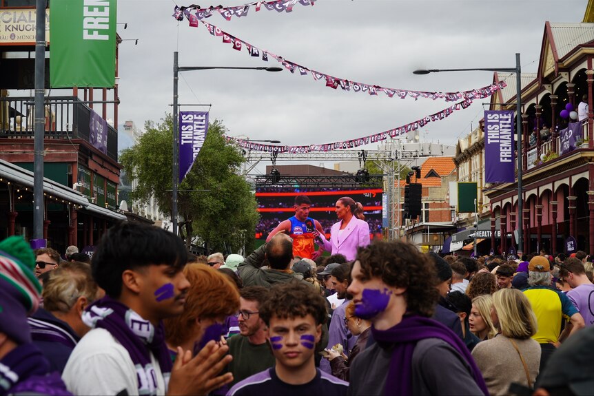 People dressed in purple with face paint walk in a street looking sad, in a big crowd.