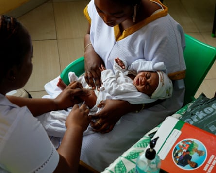 A baby held by a seated woman is injected by another woman wearing a uniform