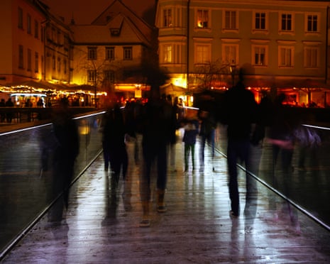 Blurred silhouettes of pedestrians walk along a wet, lit bridge at night in Ljubljana