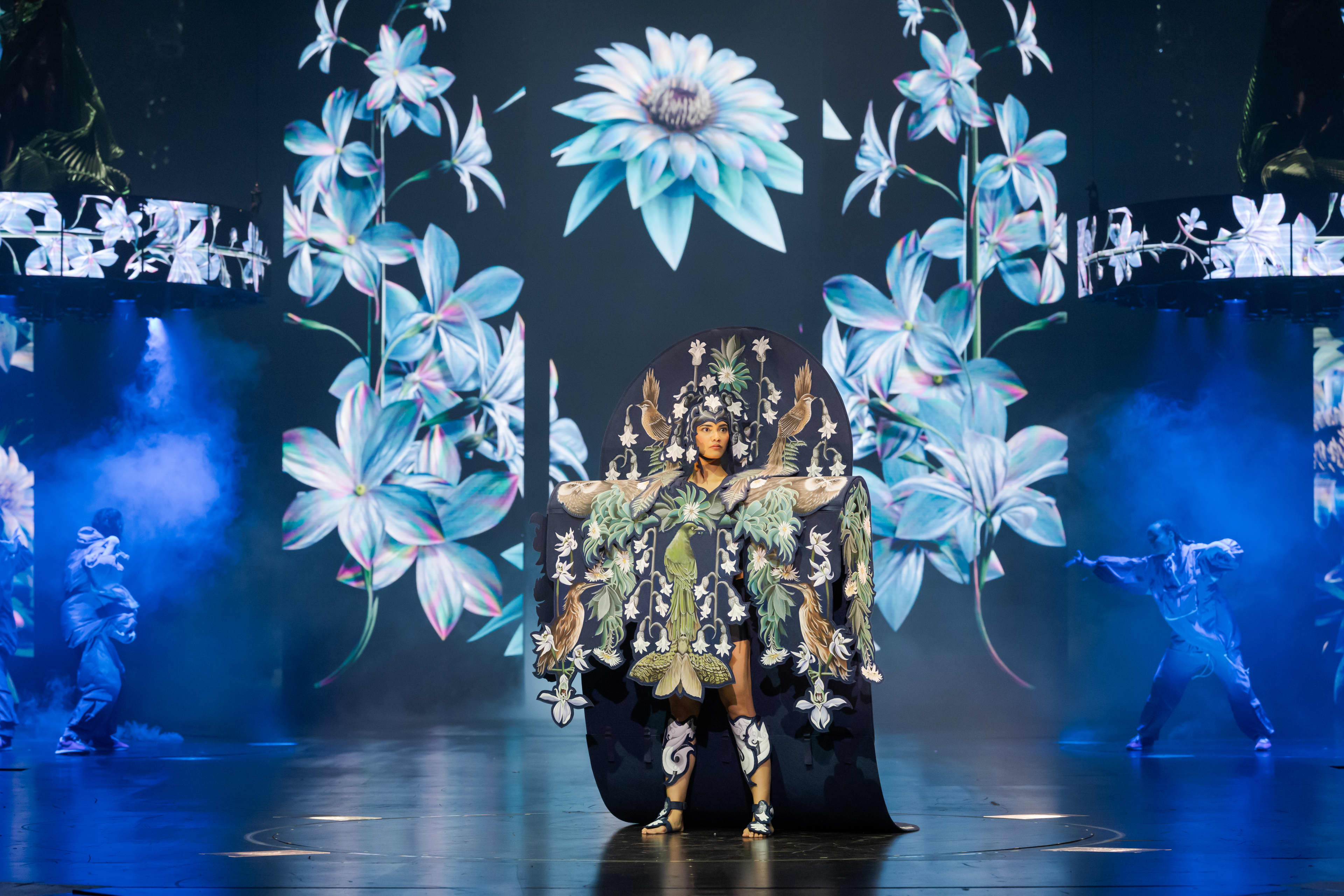 A model wears a detailed blue and white costume with headpiece decorated with feather and flower motifs.