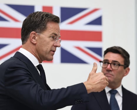 Mark Rutte gestures with his hands while Andy Burnham looks on beside him; both are standing against a white background decorated with the union flag