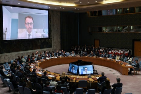 A man appears on a large screen on the wall above a horseshoe-shaped table with people sitting all around it.