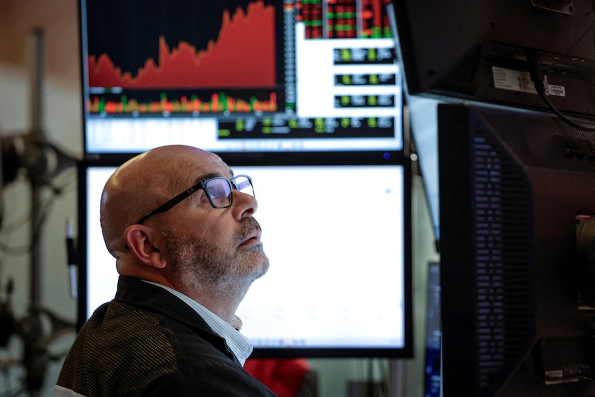 A bald middle-aged man wearing glasses sits at a desk and looks up at a computer monitor with a worried expression.