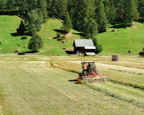 A tractor cuts hay in a Swiss meadow near a wooden cabin and forested hillside.