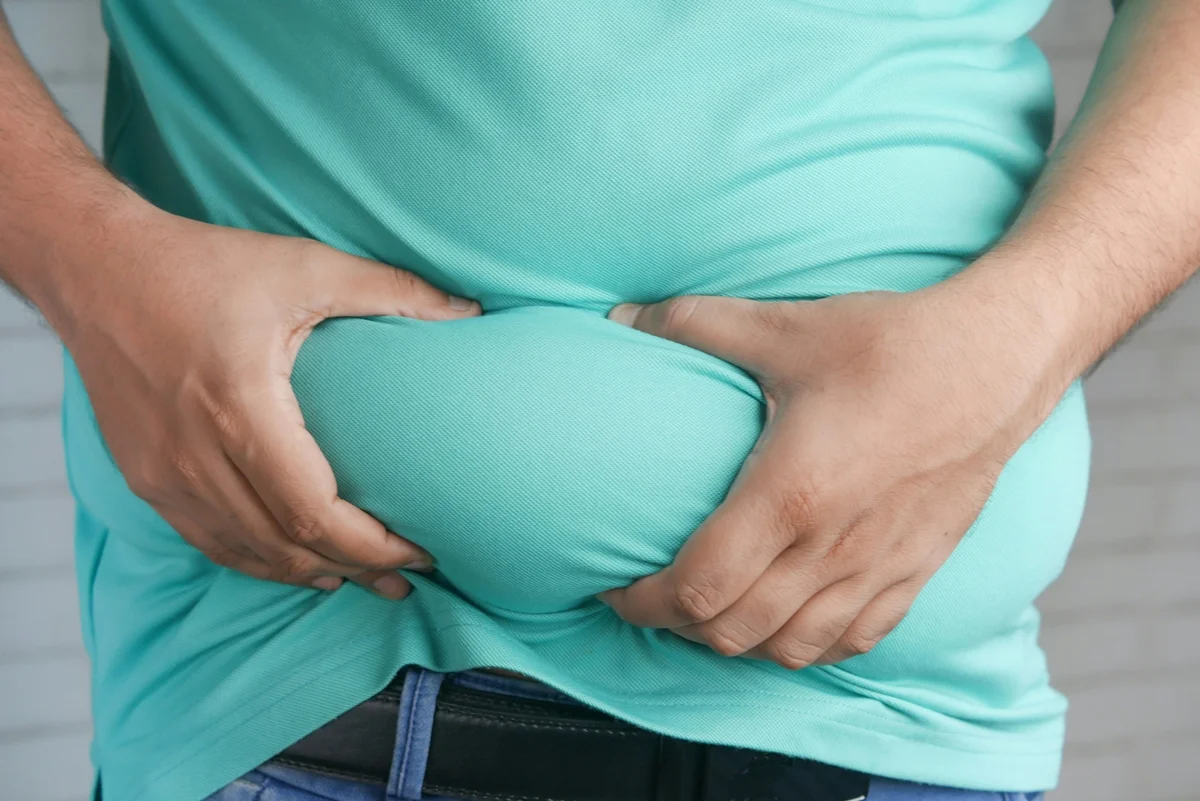 Close-up das mãos de uma pessoa segurando a própria barriga com gordura. Gordura abdominal. Metrópoles
