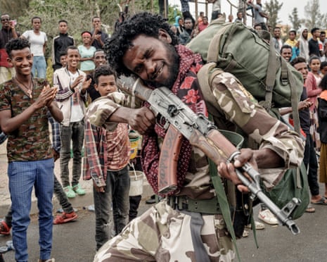 An African man in camouflage poses with an AK-47 assault rifle as bystanders applaud